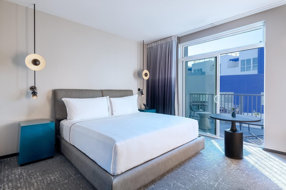 A hotel room with a bed, bedside tables, a balcony with a table and chairs, and a view of the outside.