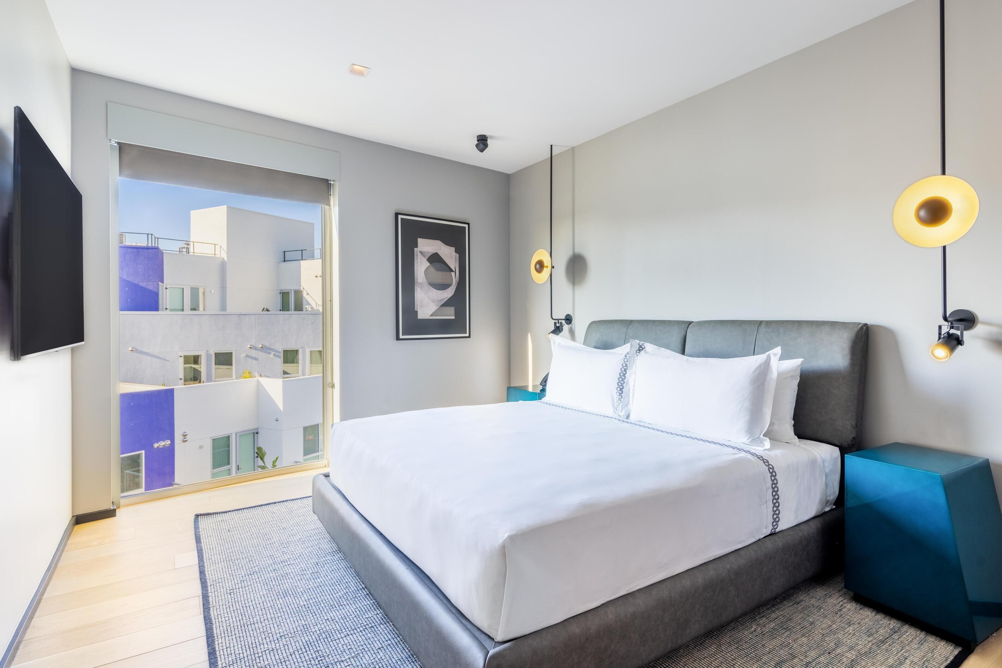 A modern hotel room with a large bed, a television, a framed artwork, and a view of the outside.