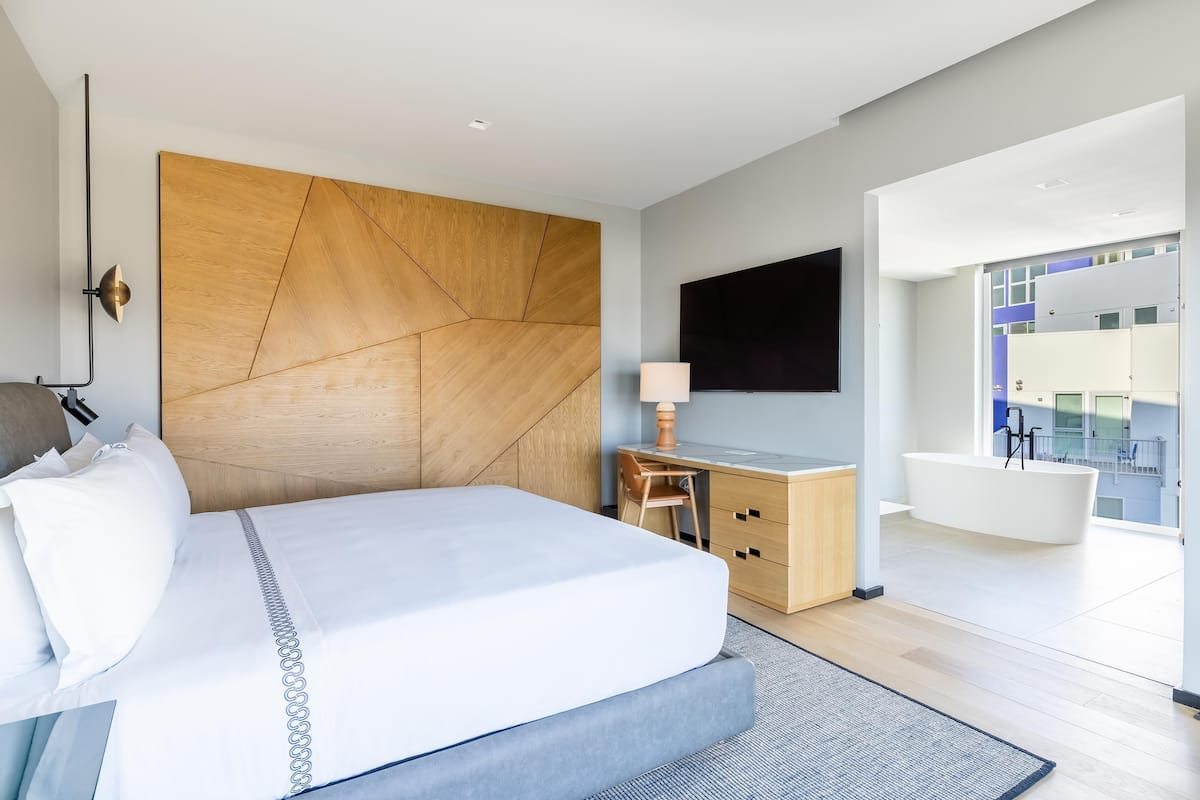 A modern hotel room with a large bed, a desk, a flat-screen TV, a bathtub, and a view of the city.