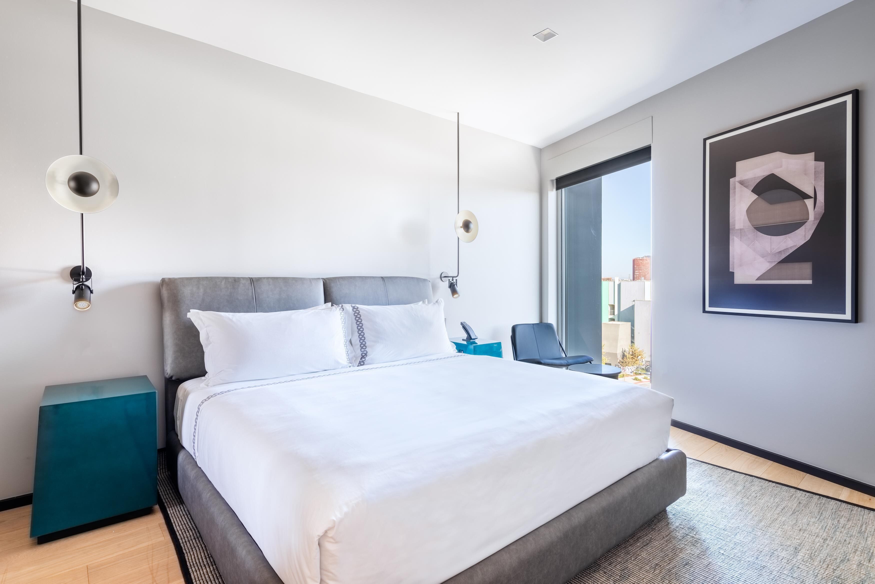 Moderni hotellihuone, jossa on suuri sänky, yöpöydät, ikkunasta avautuva näkymä ja seinällä oleva kehystetty taideteos.