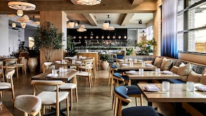 Ein Restaurant mit Holztischen und -stühlen, hängenden Pendelleuchten und einer Küche im Hintergrund.