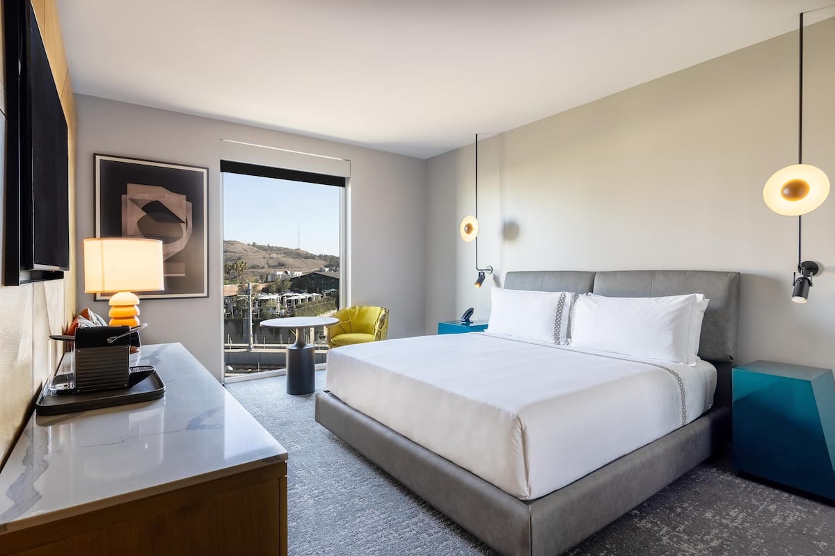 A hotel room with a bed, a desk, a chair, a nightstand, a lamp, and a view of the outside.