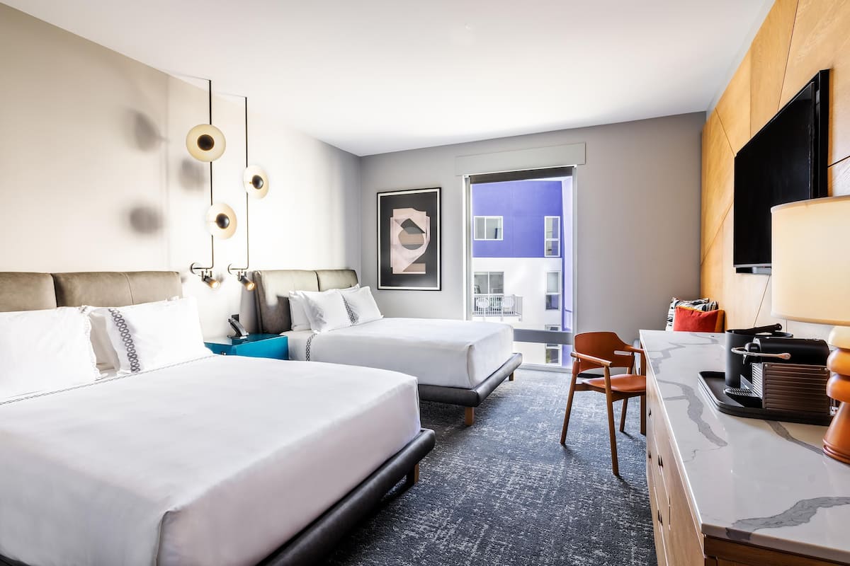 A hotel room with two beds, a desk with a lamp, a chair, a TV, and a view of a building outside.
