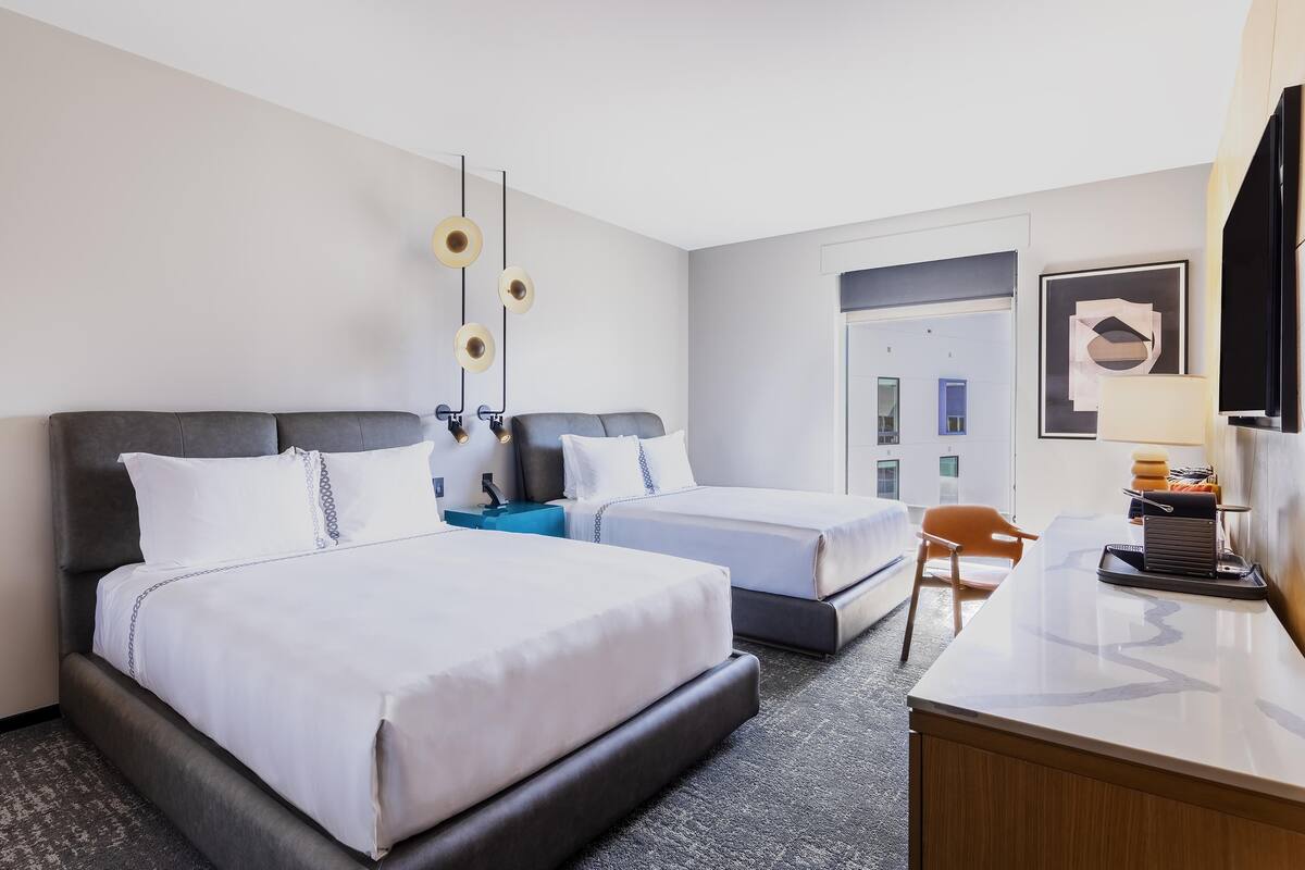 A hotel room with two beds, a desk, a chair, a TV, and a door leading to a balcony.