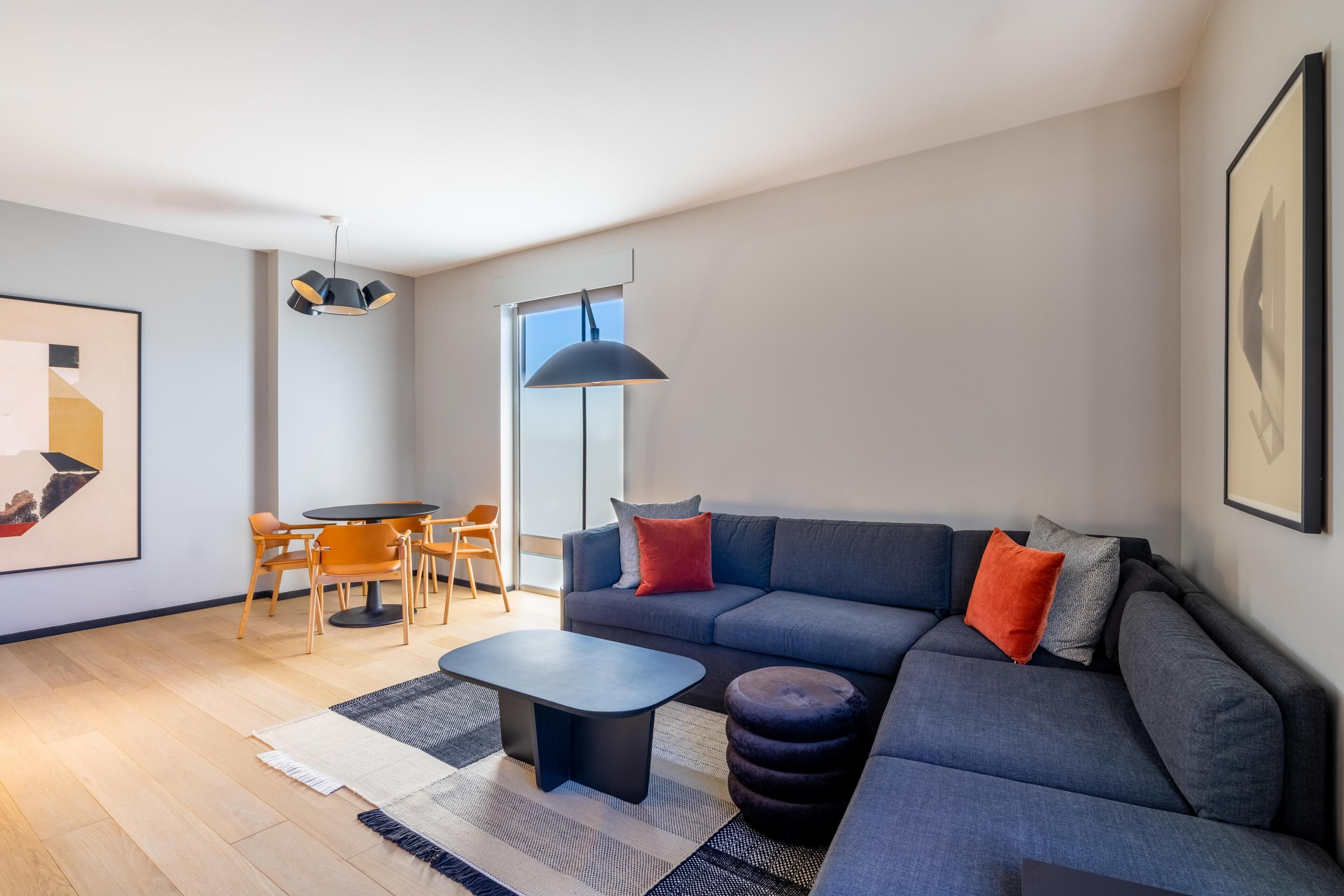 Un salon moderne comprenant un canapé d’angle, une table basse, un coin repas avec des chaises et un luminaire suspendu.