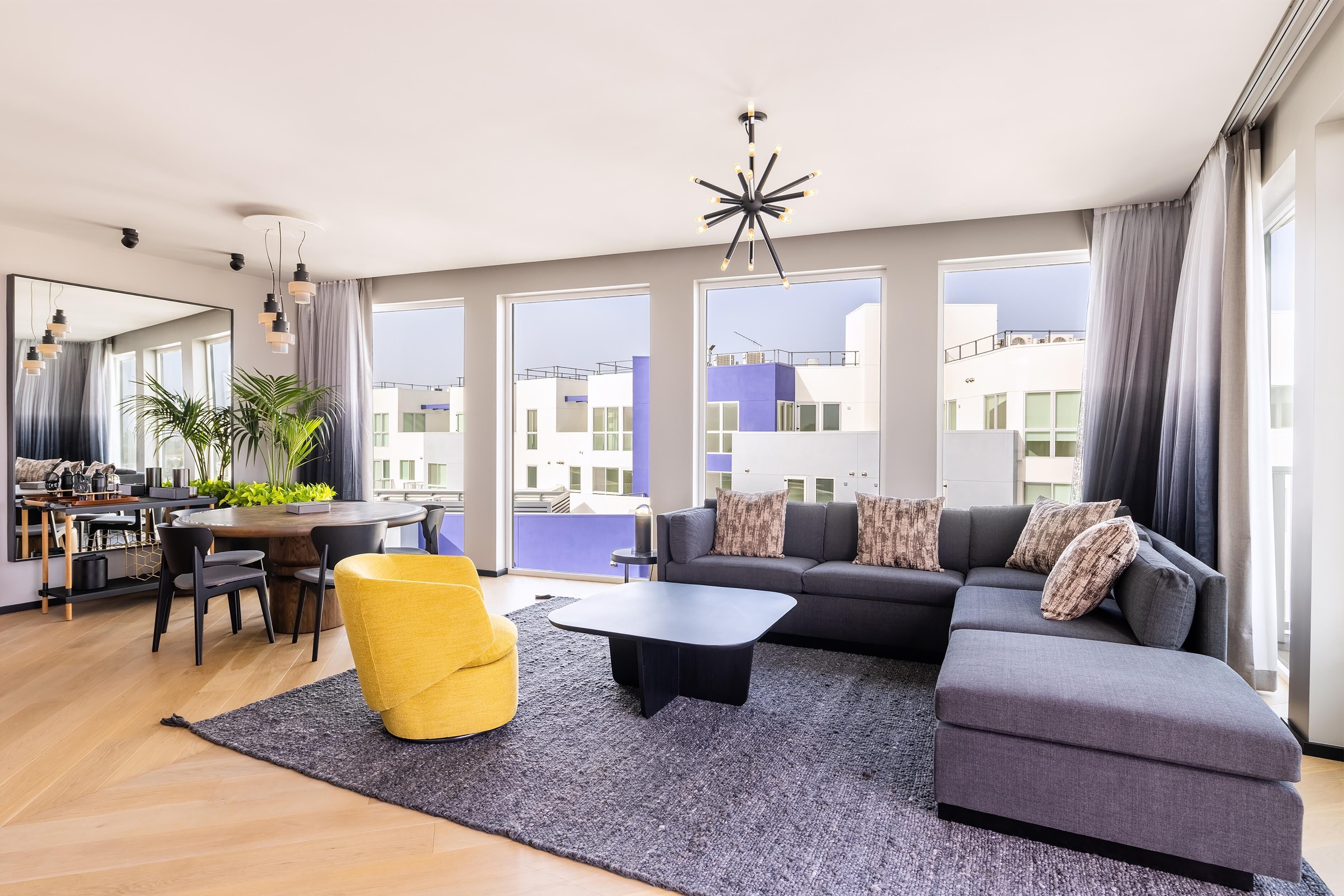 A modern living room with a sofa, coffee table, and yellow chair. There is a dining area with a table and chairs. Large windows provide views of buildings outside.
