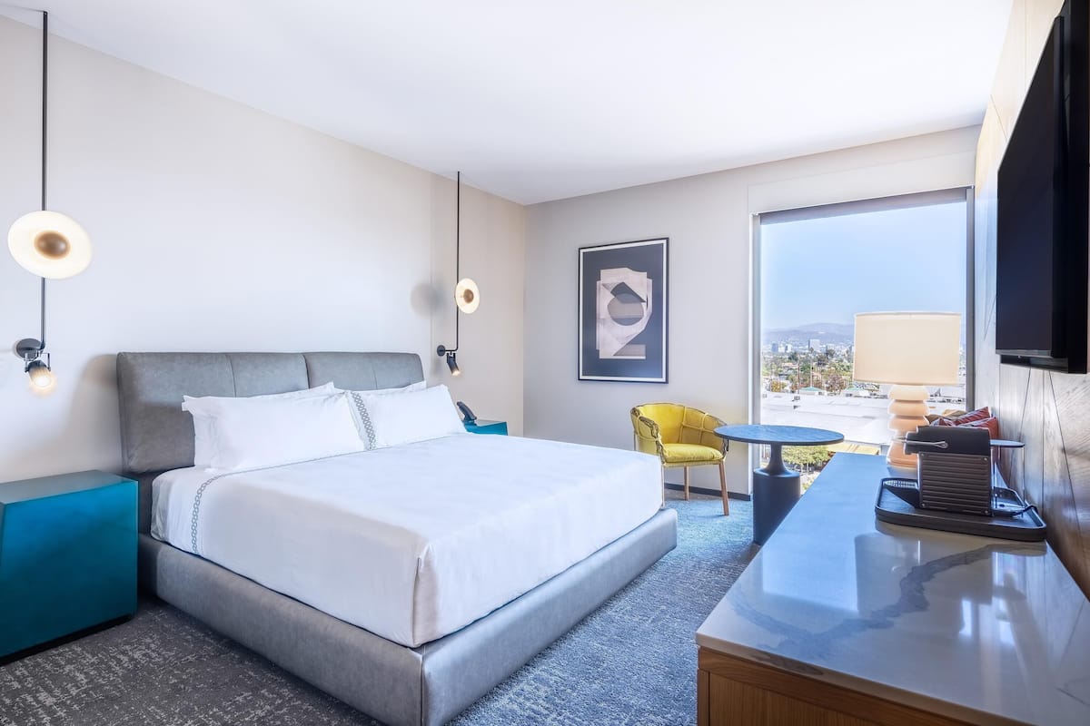 A hotel room with a bed, a desk, a chair, a TV, and a window with a city view.