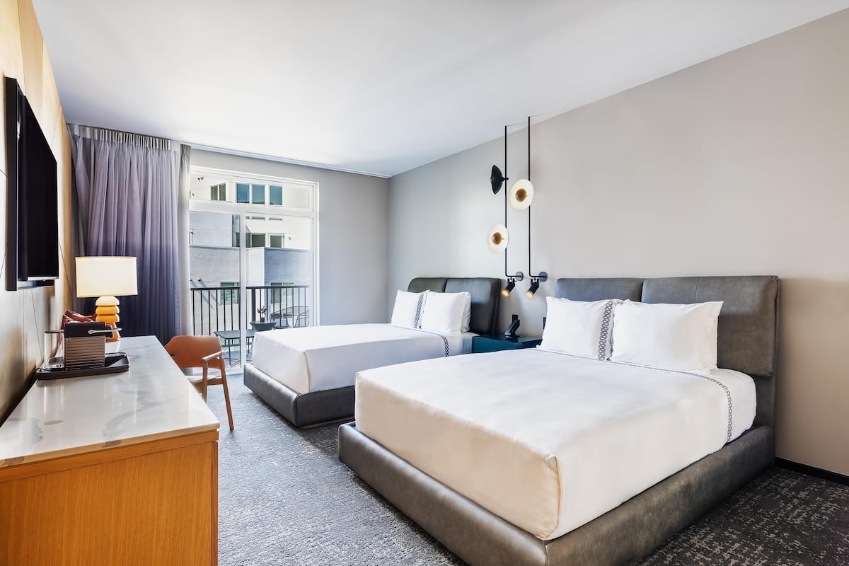 A hotel room with two beds, a desk, a chair, a TV, and a balcony with curtains.