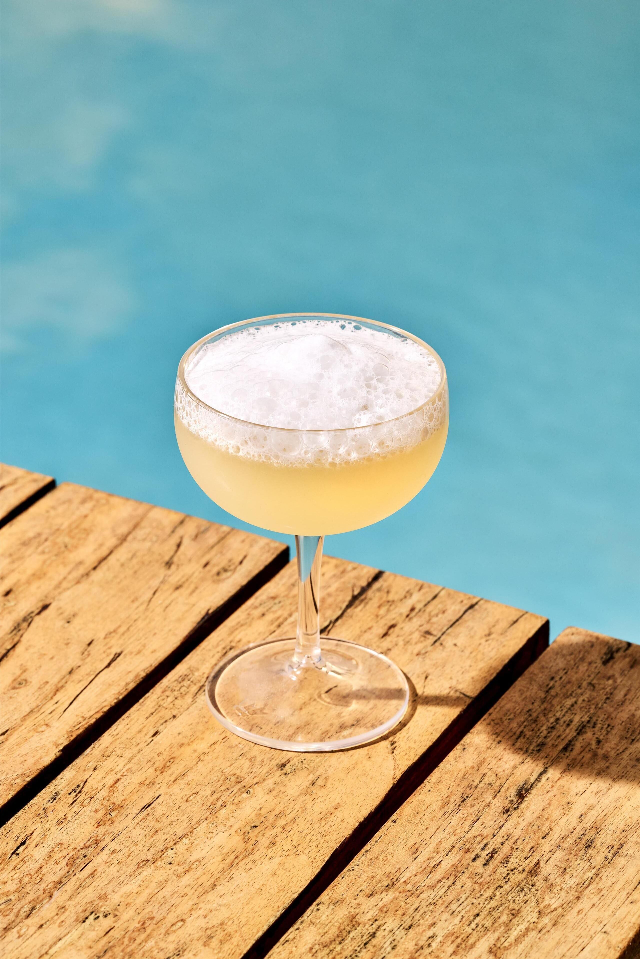 Een cocktailglas met schuim op een houten tafel bij een zwembad.