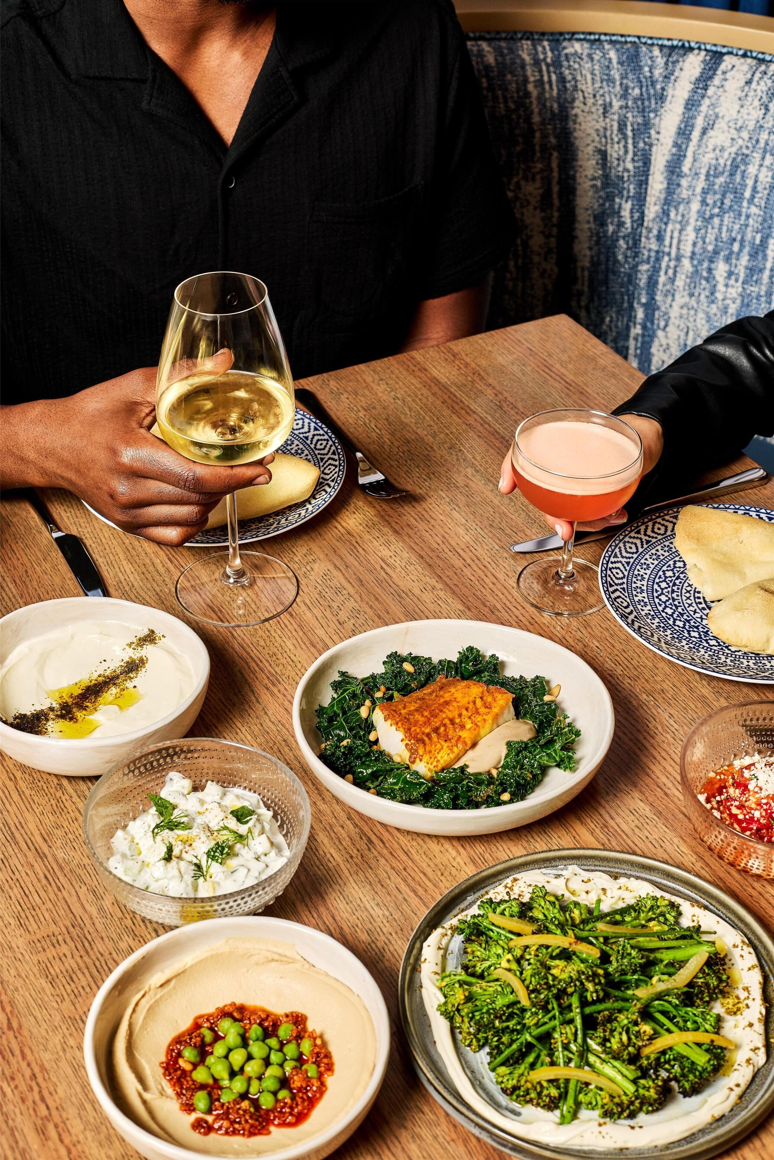 Une table à manger avec différents plats et boissons, une personne tenant un verre de vin blanc, et un coussin bleu à motifs en arrière-plan.