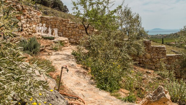Un sendero de piedra serpentea por un paisaje natural, salpicado de flores amarillas y flanqueado por una pared de piedra con estatuas.