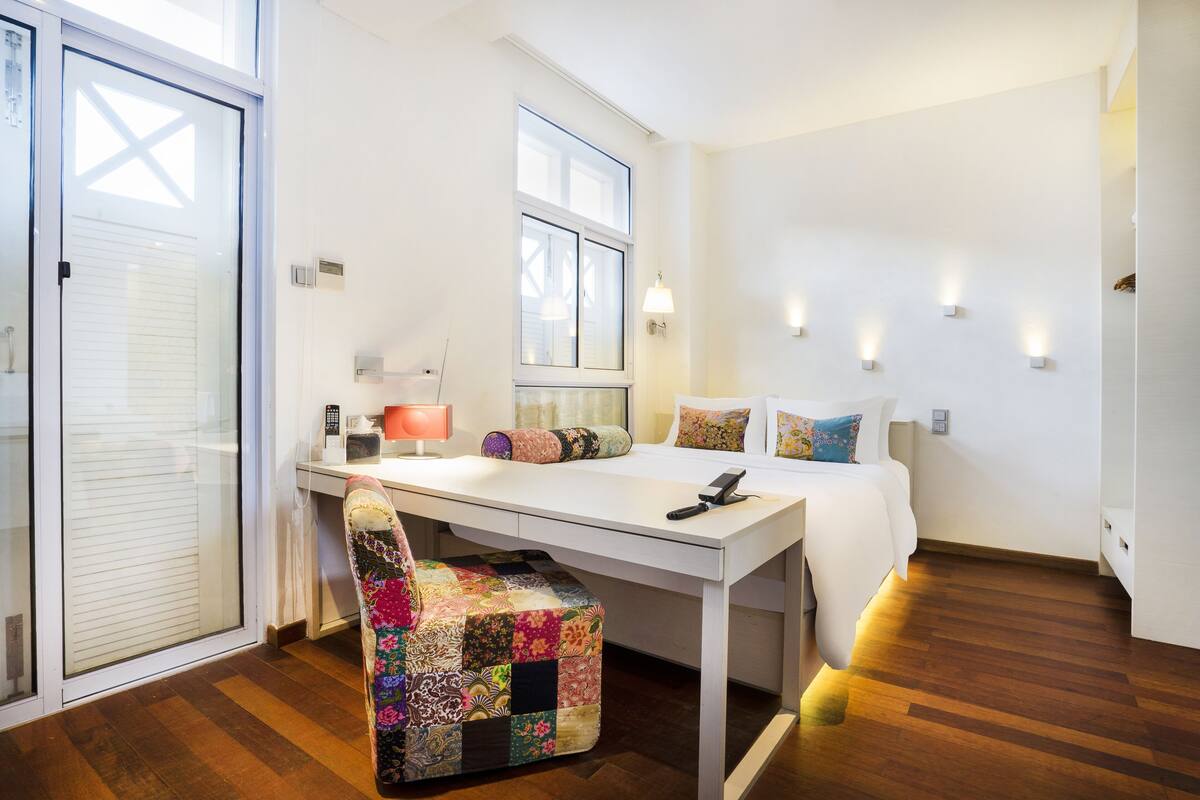 Une chambre moderne avec un bureau blanc, un fauteuil coloré à motifs matelassés, un lit avec de nombreux coussins et des lampes fixées au mur.