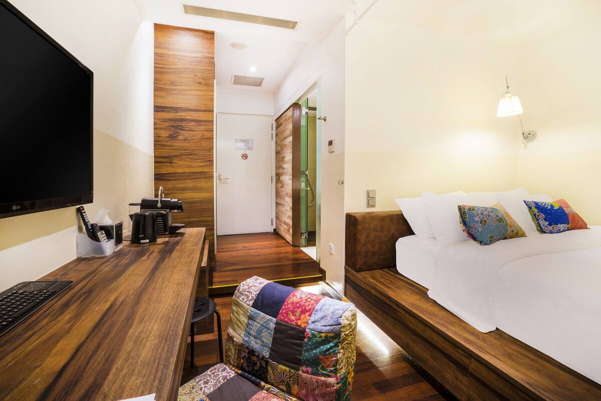 Une chambre d’hôtel avec un bureau en bois, un lit agrémenté d’oreillers colorés, une couverture patchwork et un téléviseur fixé au mur.