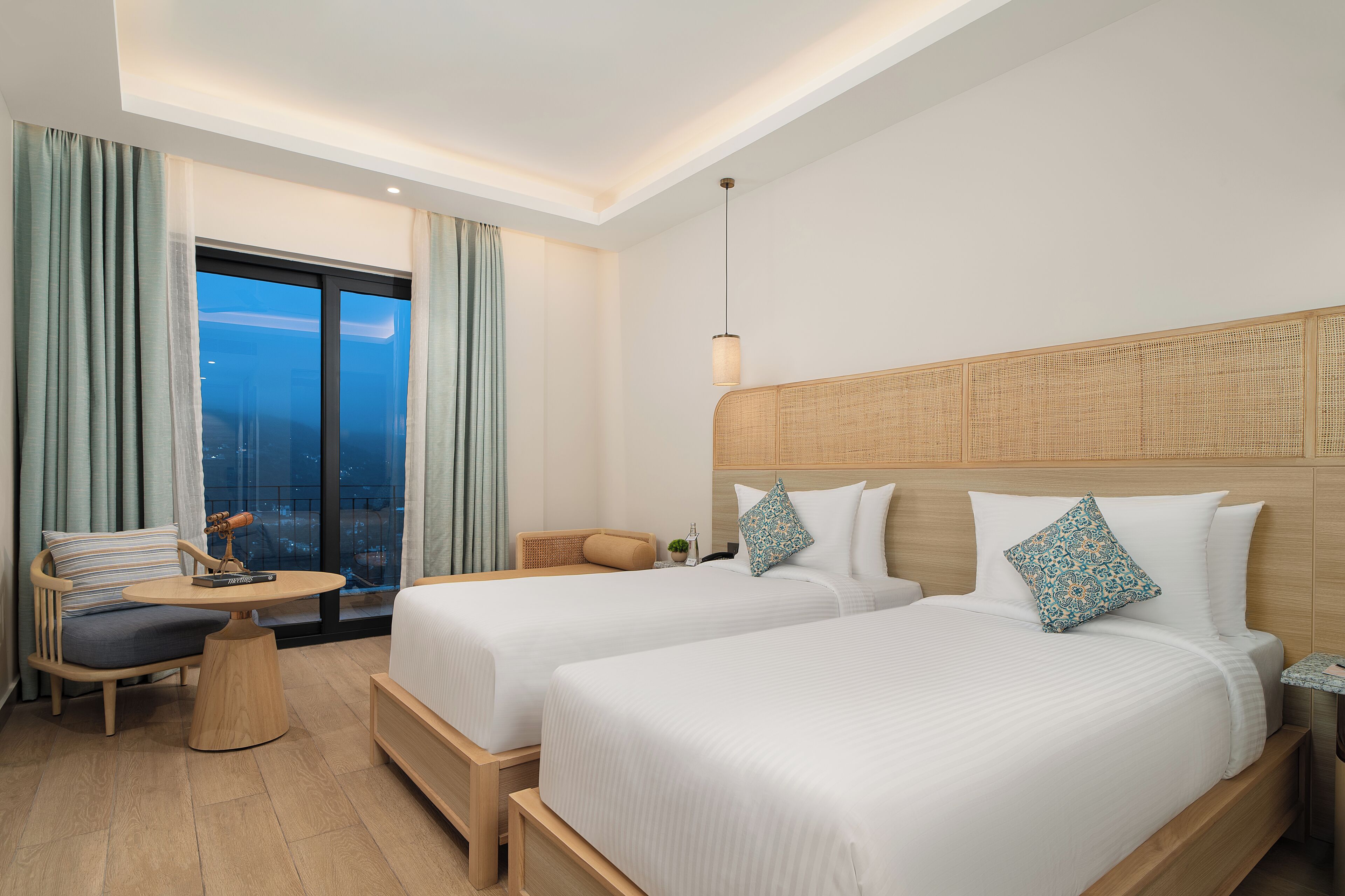 A hotel room with two beds, a small table, a chair, and a view of the city.