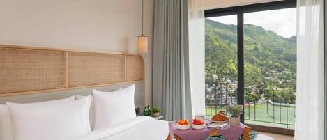 Habitación de hotel con una cama grande, vistas a un paisaje montañoso y una mesa con alimentos para el desayuno.