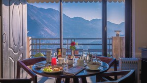 A dining table set with breakfast items, a vase with a flower, and a view of mountains through large windows. - Rosewood Retreat Bhimtal (Nainital)