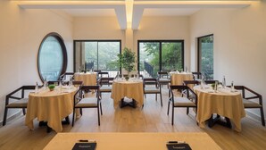 A dining area with round tables, chairs, and a view of a mountainous landscape through large windows. - Rosewood Retreat Bhimtal (Nainital)