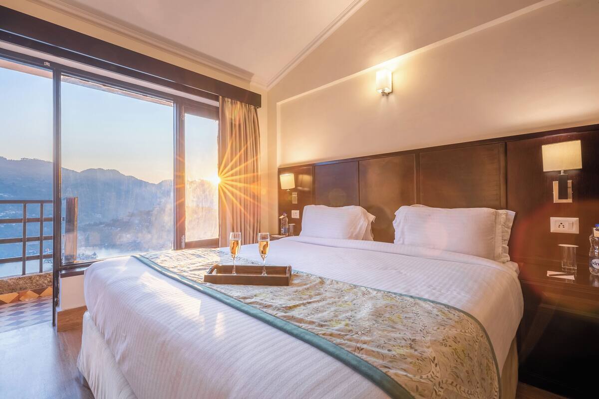 Habitación de hotel con una cama grande, una mesita de noche, una lámpara y vistas a las montañas a través de la ventana.