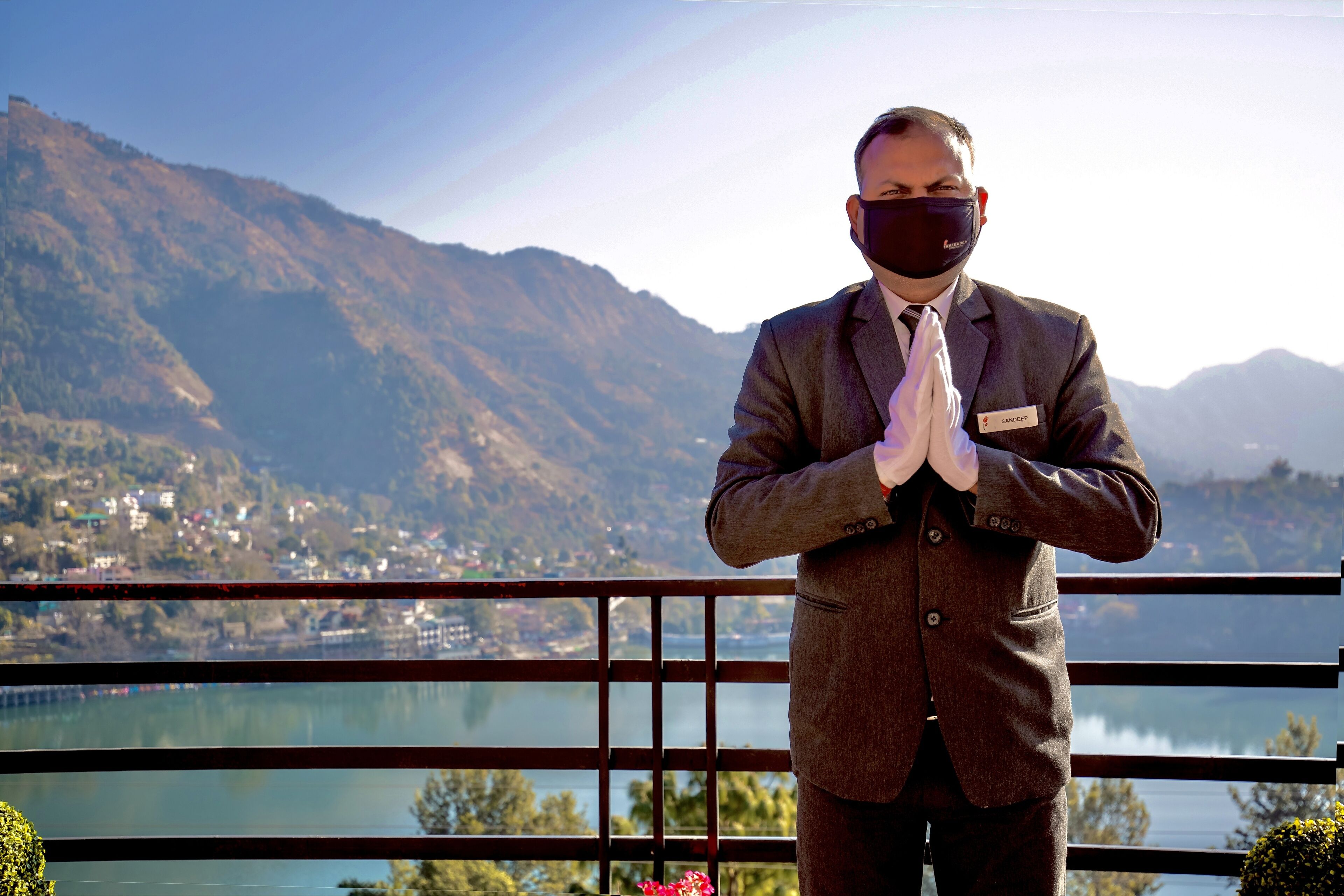 Un hombre con traje de pie, con las manos juntas, observa un paisaje montañoso con un lago y un pueblo a lo lejos.