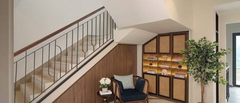 A staircase with a wooden handrail and a glass railing, a dark upholstered chair, a small table with a vase of flowers, a bookshelf with books and decorative items, and a potted plant.