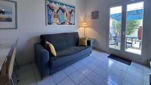 Una habitación con sofá azul, un cuadro de casas coloridas, una lámpara y una puerta que da a un espacio exterior con mesas y sombrillas.