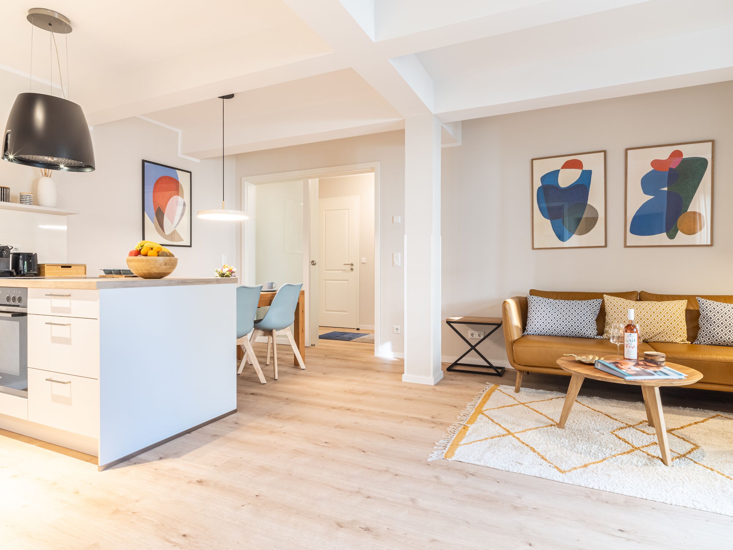 Een moderne keuken met een centraal eiland, een eethoek met stoelen, een bank met opvallende kussens en abstracte muurkunst.