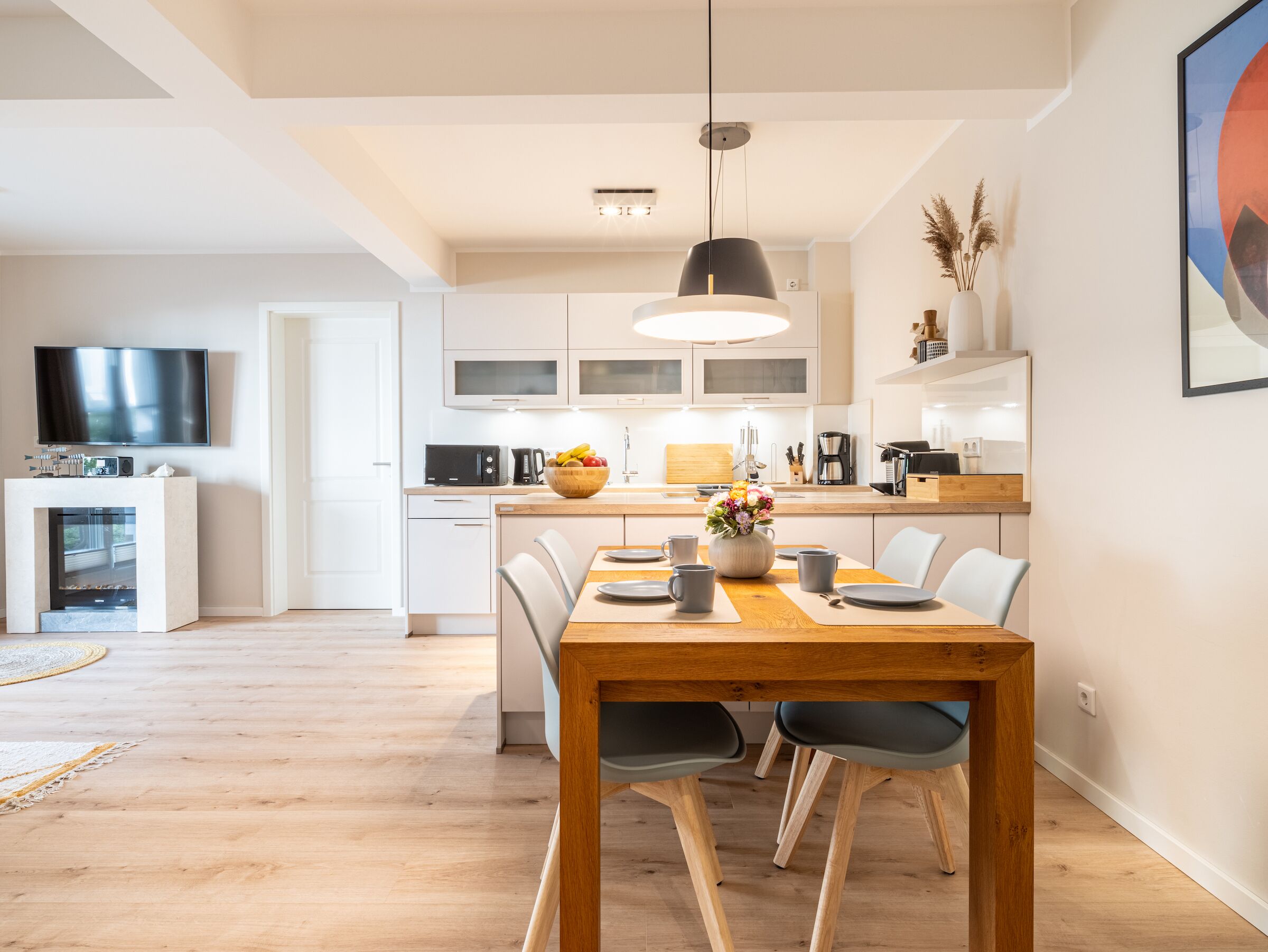 A modern kitchen with white cabinets, a wooden dining table, and a TV mounted on the wall.