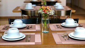 A dining table set for four with white cups, saucers, and cutlery on pink placemats. A vase with flowers is centered on the table. - Hotel Belvedere Araras - Fácil Acesso Hospital São Leopoldo e Faculdades (Araras)