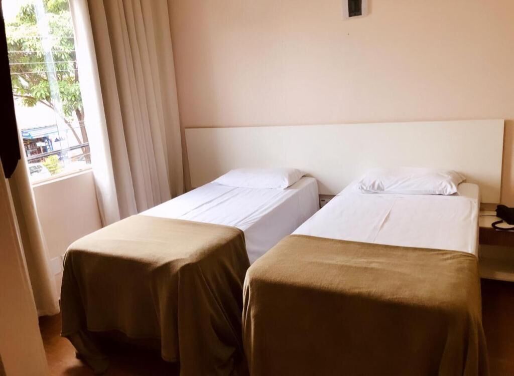 Quarto de hotel com duas camas de solteiro, uma janela com cortinas e uma mesinha com telefone.