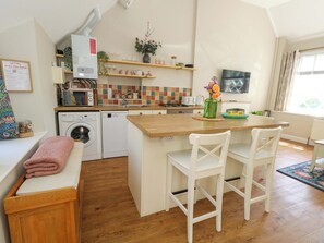 A compact kitchen with a washing machine, microwave, and a small dining area with two chairs. - Sea Pink Cottage (Swanage)