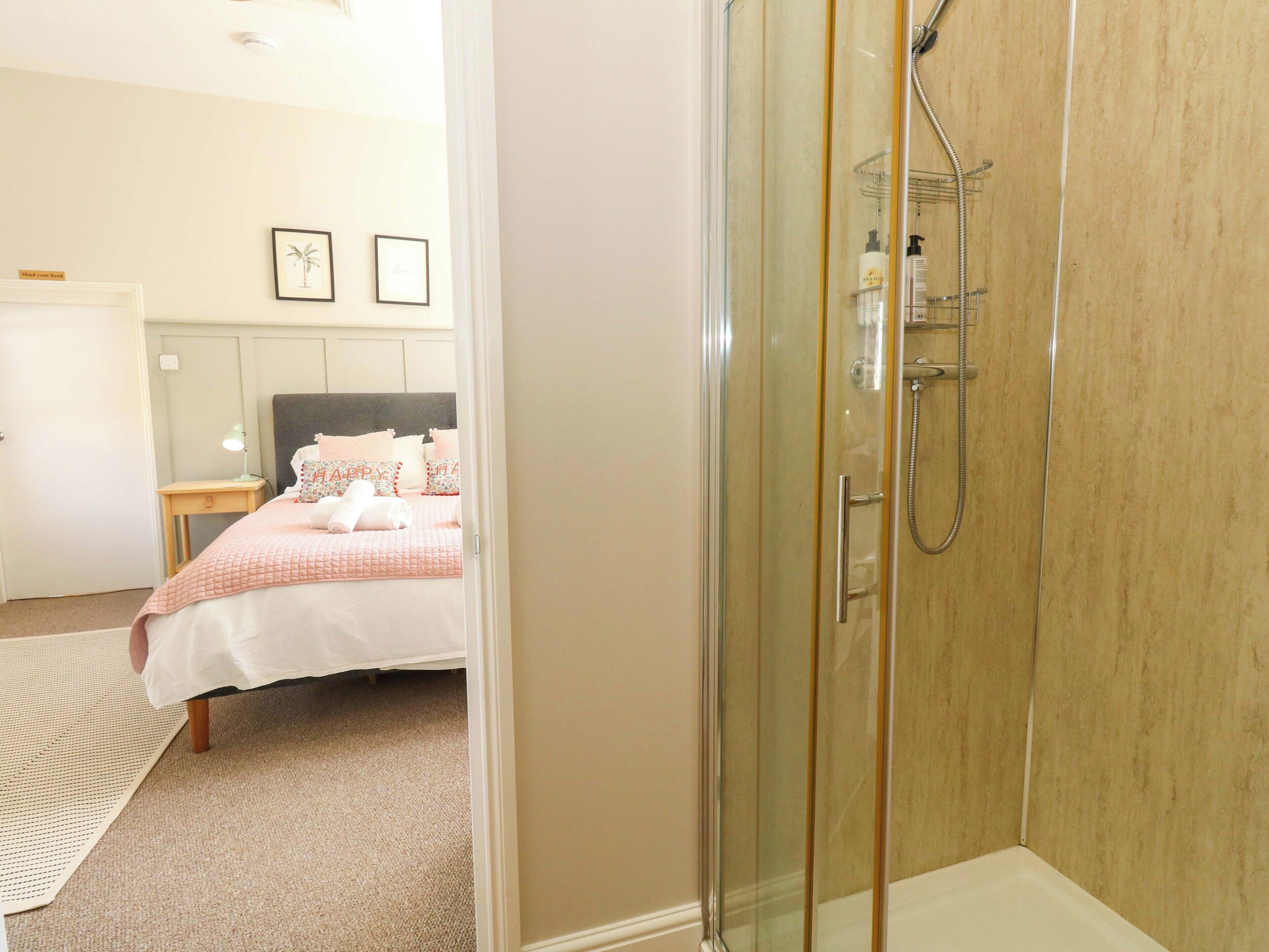 A hotel room with a glass-enclosed shower, a bed with a pink bedspread, and a small bedside table with a lamp.