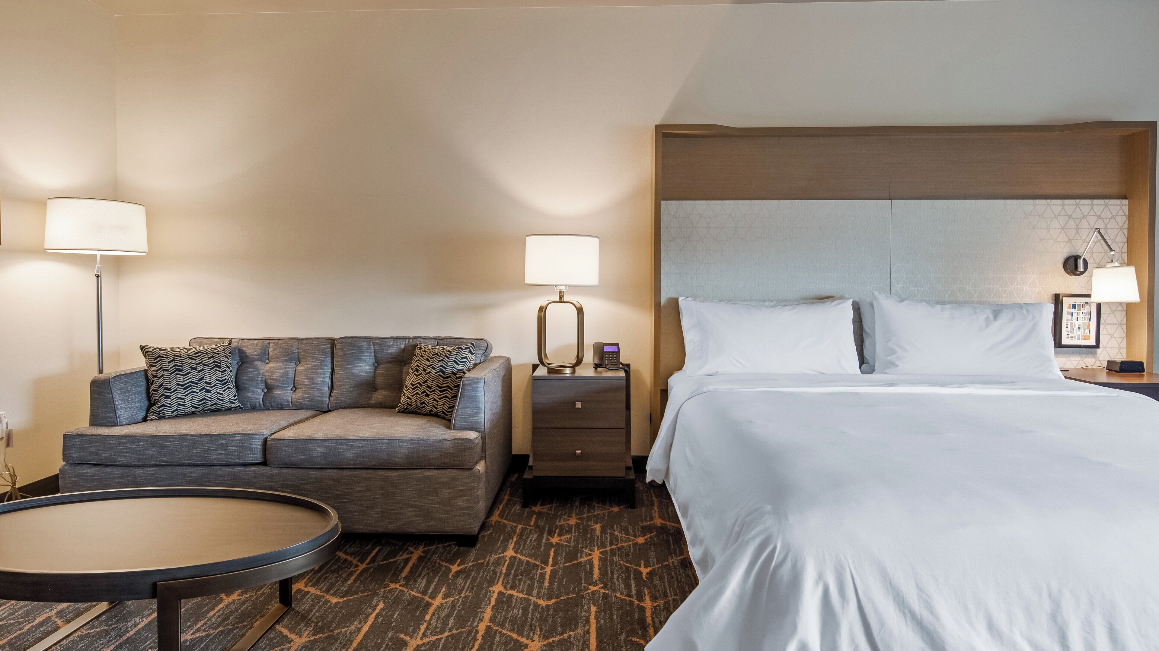 A hotel room with a bed, a sofa, a nightstand, a lamp, and a small round table.