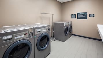 A laundry facility with two front-loading washing machines and a dryer, a metal rack, and three informational signs on the wall.