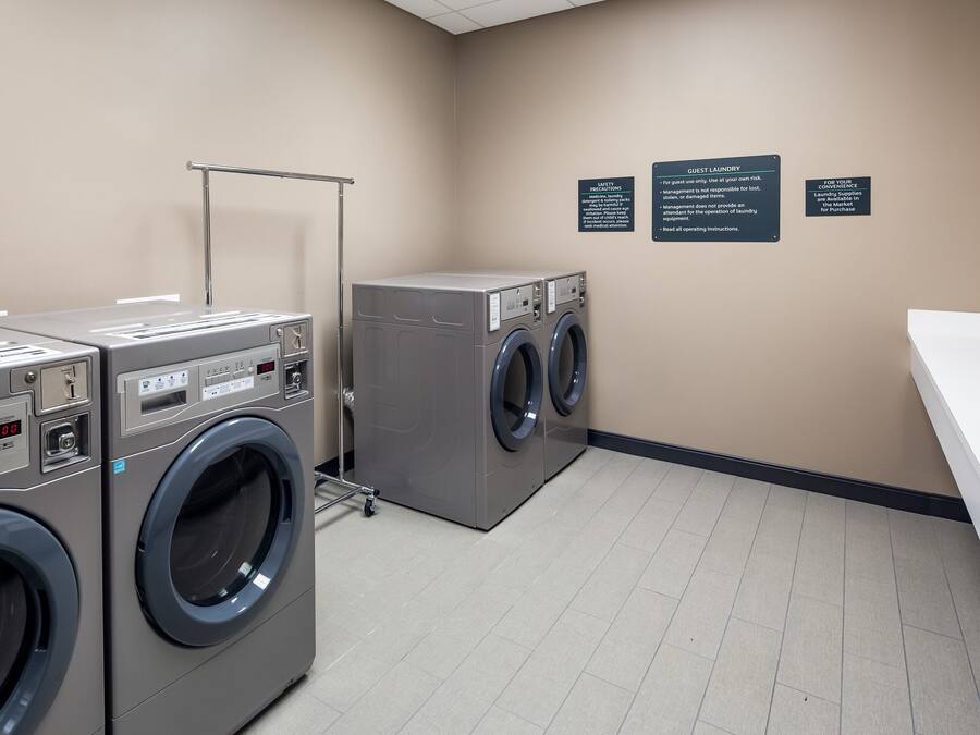 A laundry facility with two front-loading washing machines and a dryer, a metal rack, and three informational signs on the wall.