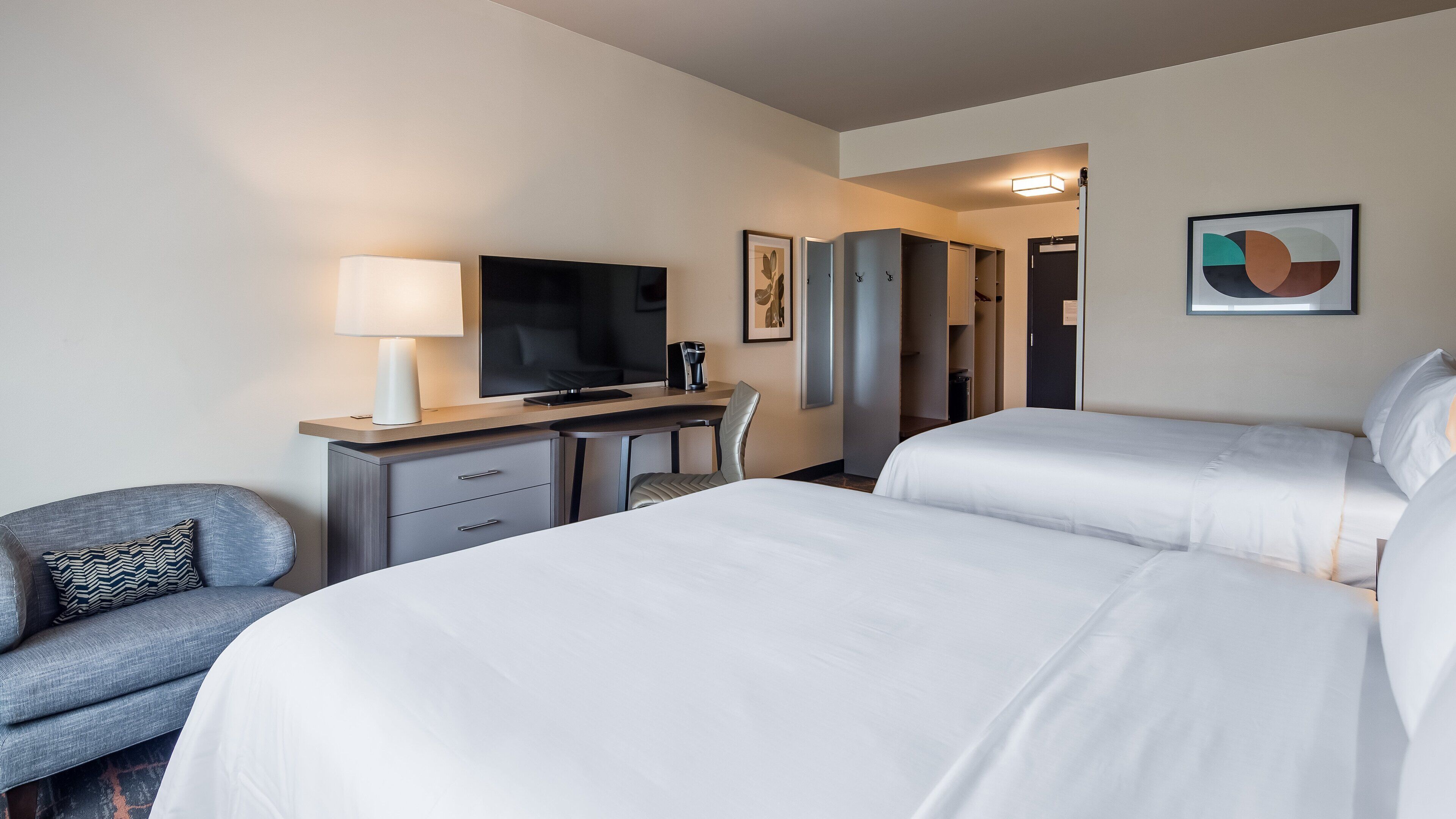 A hotel room with two beds, a desk with a TV, a chair, and a lamp.