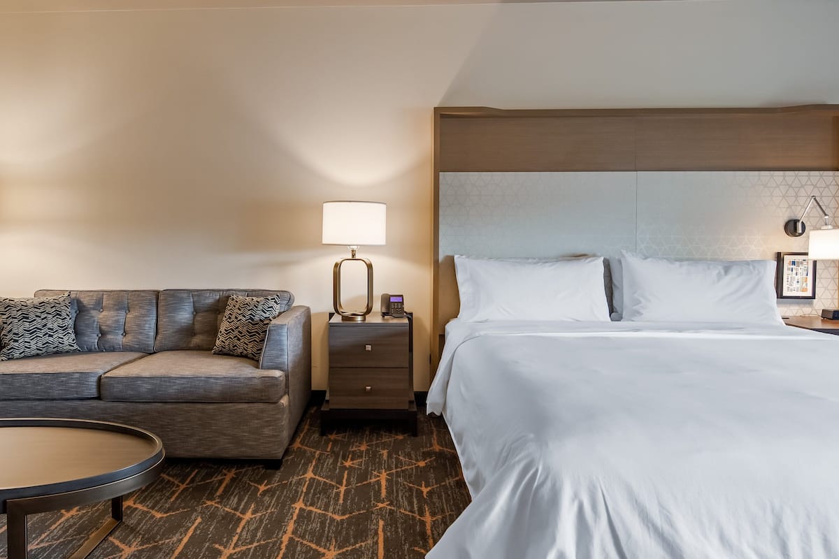 Habitación de hotel con cama, sofá, mesita de noche, lámpara y una pequeña mesa redonda.