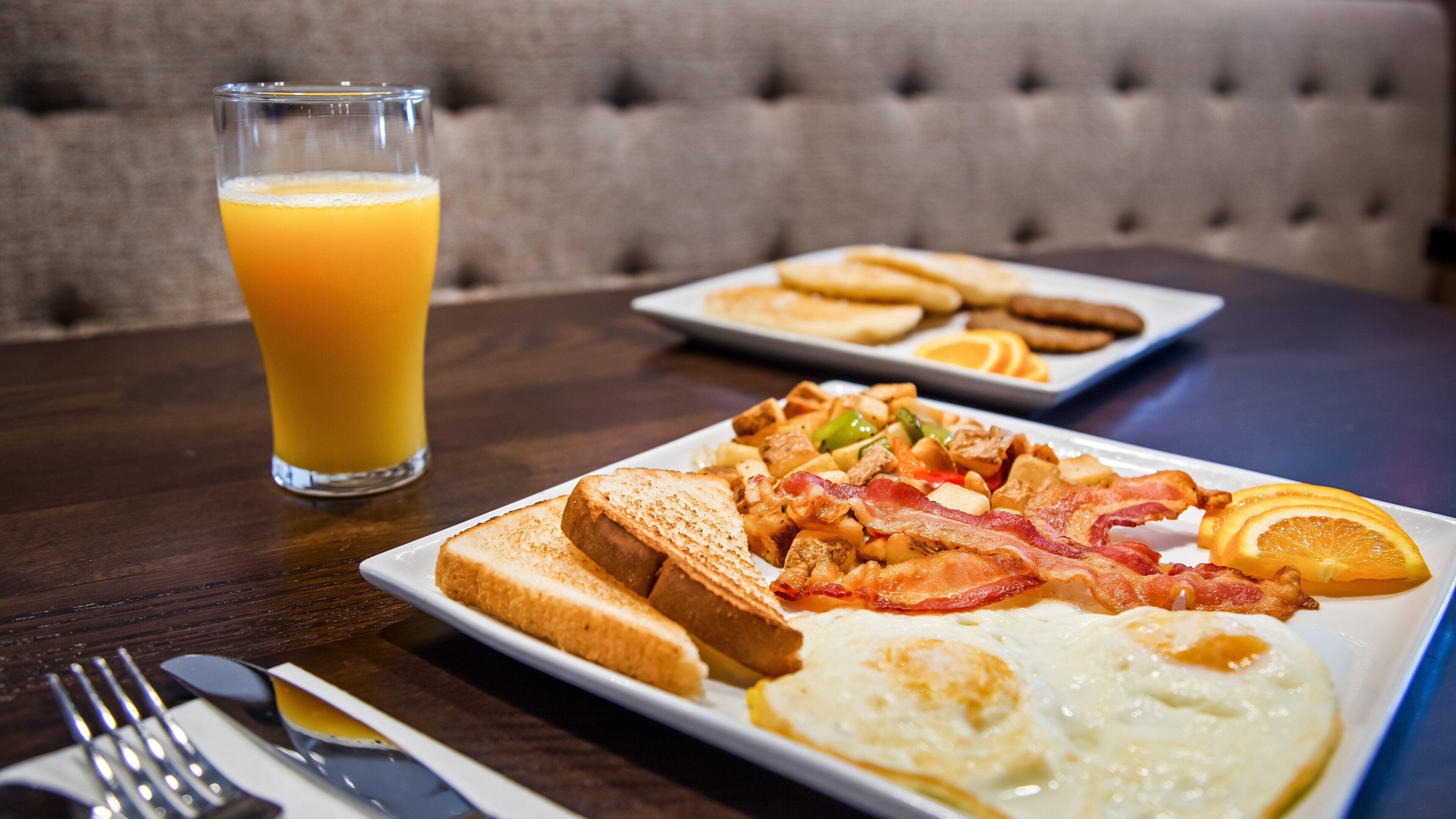 A breakfast plate with toast, bacon, eggs, and orange slices, and a glass of orange juice on a wooden table.