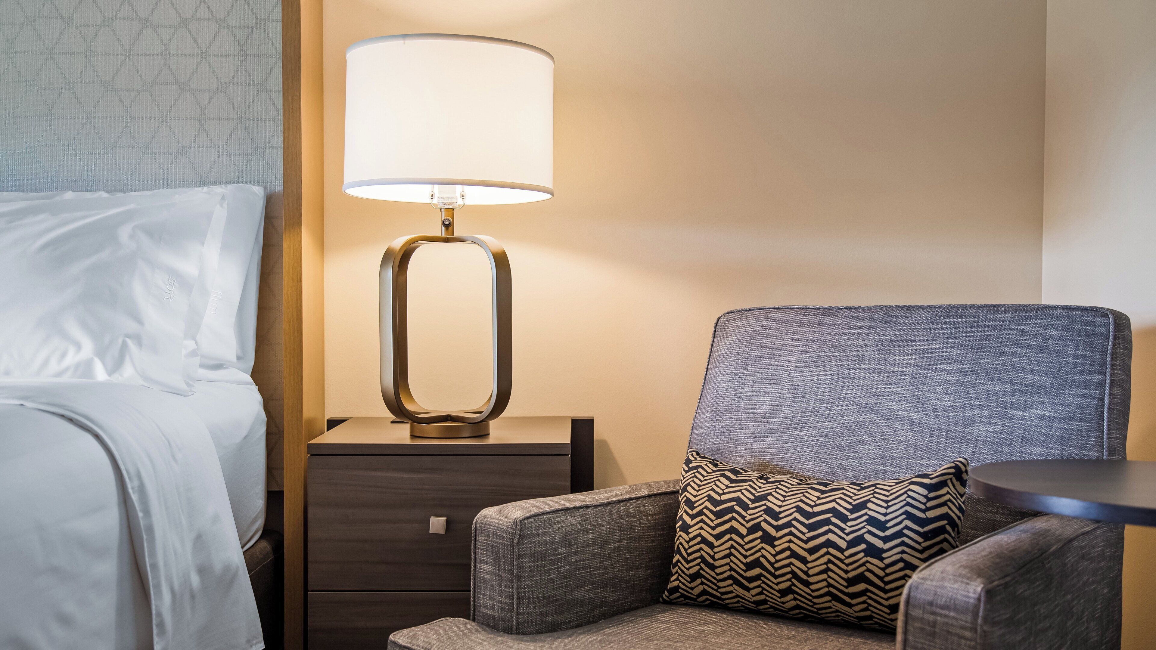 A hotel room with a bed, a nightstand with a lamp, a grey upholstered armchair, and a small table.