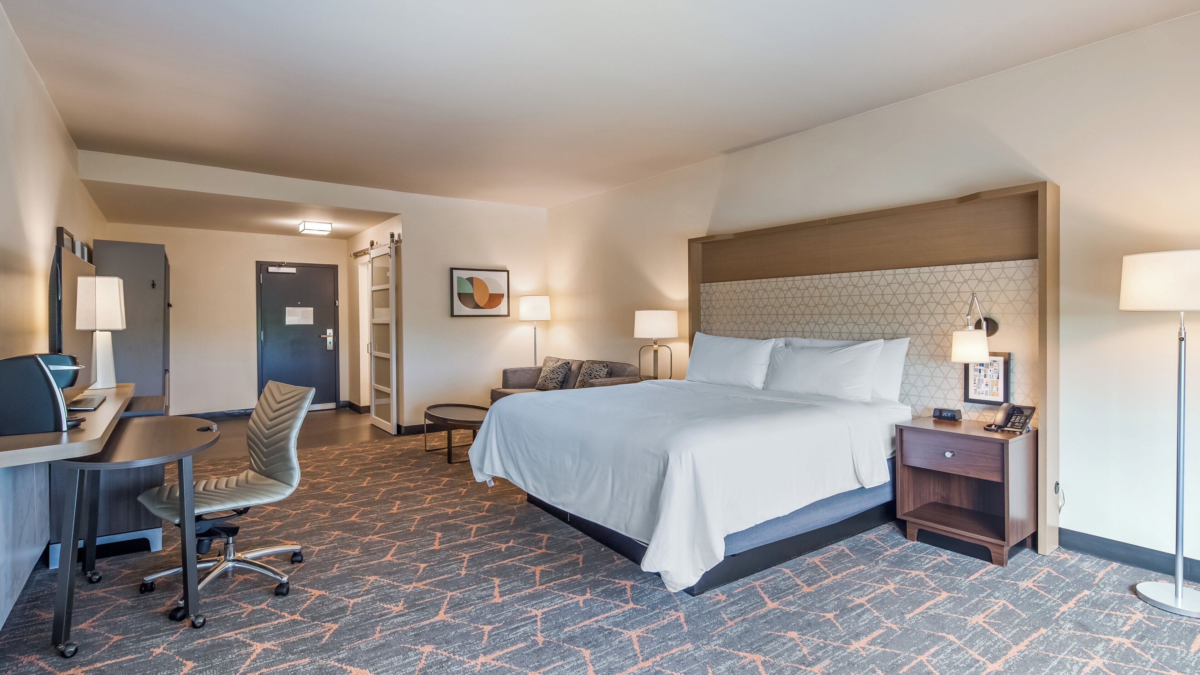 Quarto de hotel com cama, escrivaninha, cadeira e uma pequena mesa com abajur.