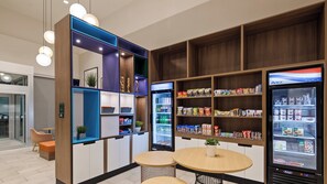 Uma kitchenette moderna com eletrodomésticos embutidos, uma geladeira repleta de lanches e uma área de estar com duas mesas redondas.