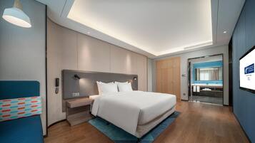 A modern hotel room with a large bed, a bedside table, a desk, and a bathroom visible through an open door.