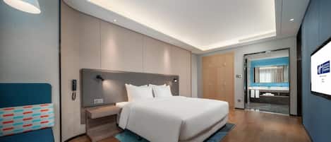 A modern hotel room with a large bed, a bedside table, a desk, and a bathroom visible through an open door.