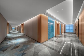 Ein moderner Hotelkorridor mit einem geometrisch gemusterten Boden, eingelassener Beleuchtung und Blick zum Himmel durch eine Glastür.