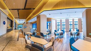A modern hotel lobby with a central reception desk, seating areas, and a view of the dining area.