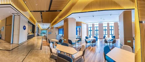 A modern hotel lobby with a central reception desk, seating areas, and a view of the dining area.