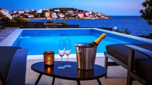 Escena a la piscina amb vista a una vila costanera, una taula amb dues copes i una ampolla de xampany, i una espelma decorativa.