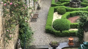 A courtyard with a stone wall, a bench, a statue, and a well-maintained garden with hedges and a fountain.