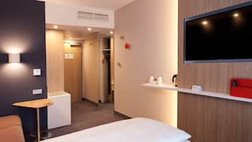 A hotel room with a bed, a small table, a TV, a shelf with a kettle and a red phone, and a closet.