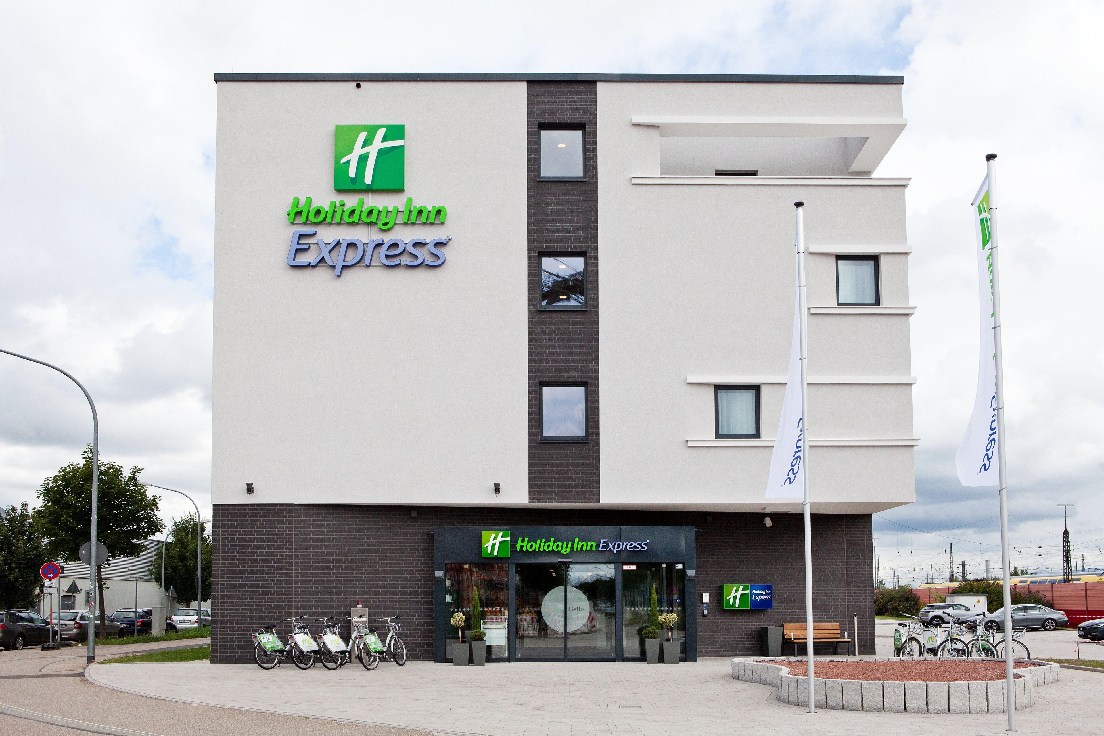 Een Holiday Inn Express gebouw met een modern ontwerp, herkenbaar aan een groot logo, een brede entree en een parkeerplaats met fietsen.