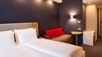 A hotel room with two beds, a red chair, a small table, and a wardrobe.